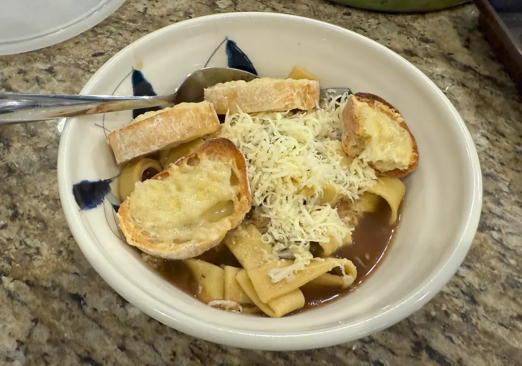 Bowl of French onion beef and noodles topped with cheesy Gruyère bread – slow braised chuck roast with caramelized onions and pasta in rich broth