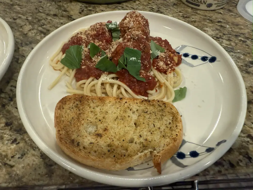 A plate of spaghetti topped with tender beef, veal, and pork meatballs simmered in a rich tomato and shallot sauce. Fresh basil and grated Parmesan are scattered over the top, with steam rising from the glossy, deep-red sauce.