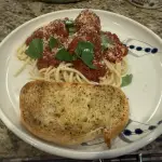 A plate of spaghetti topped with tender beef, veal, and pork meatballs simmered in a rich tomato and shallot sauce. Fresh basil and grated Parmesan are scattered over the top, with steam rising from the glossy, deep-red sauce.
