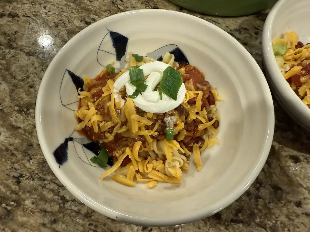 A steaming bowl of Jeff’s Fiery Chili topped with melted cheddar, sliced green onions, and a dollop of sour cream — rich, smoky red color with visible bits of beef, sausage, and peppers.