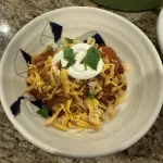 A steaming bowl of Jeff’s Fiery Chili topped with melted cheddar, sliced green onions, and a dollop of sour cream — rich, smoky red color with visible bits of beef, sausage, and peppers.