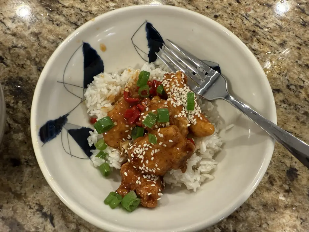 Homemade orange chicken glazed in a glossy orange sauce with sliced Fresno chilies and orange zest on top, served over jasmine rice in a white bowl.