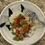 Homemade orange chicken glazed in a glossy orange sauce with sliced Fresno chilies and orange zest on top, served over jasmine rice in a white bowl.