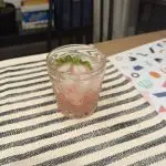 A glass of pale pink homemade raspberry lemonade over ice with a few mint leaves floating on top, sitting on a kitchen counter in soft natural light.