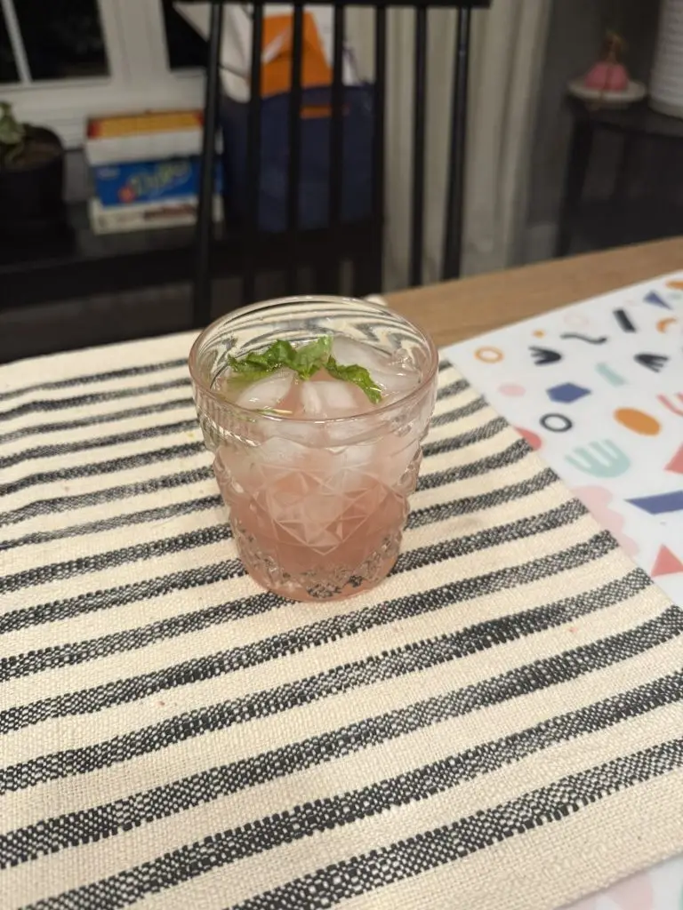 A glass of pale pink homemade raspberry lemonade over ice with a few mint leaves floating on top, sitting on a kitchen counter in soft natural light.