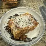 A golden-brown lasagna just out of the oven, layers of rich red meat sauce and creamy white béchamel visible beneath melted cheese, finished with fresh chives and a drizzle of olive oil.