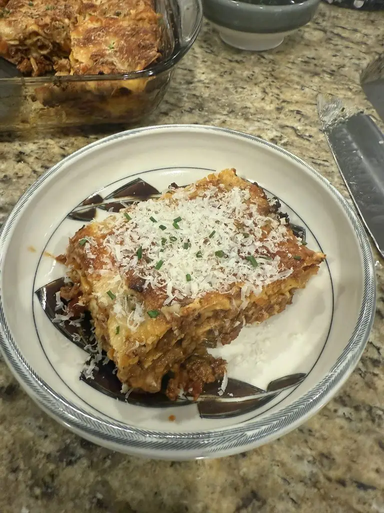 A golden-brown lasagna just out of the oven, layers of rich red meat sauce and creamy white béchamel visible beneath melted cheese, finished with fresh chives and a drizzle of olive oil.