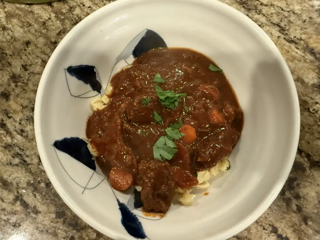 A bowl of Hungarian goulash served over buttery spätzle. Tender chunks of paprika-braised beef sit in a rich reddish sauce with carrots and peppers, topped with fresh parsley.
