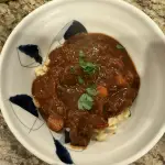 A bowl of Hungarian goulash served over buttery spätzle. Tender chunks of paprika-braised beef sit in a rich reddish sauce with carrots and peppers, topped with fresh parsley.