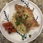 A plate of chicken piccata served over angel hair pasta with a lemon-butter-caper sauce. The chicken is golden and glossy, topped with capers and fresh parsley. On the side are blistered cherry tomatoes, lightly sautéed in butter and garlic. The dish looks bright, fresh, and restaurant-quality.