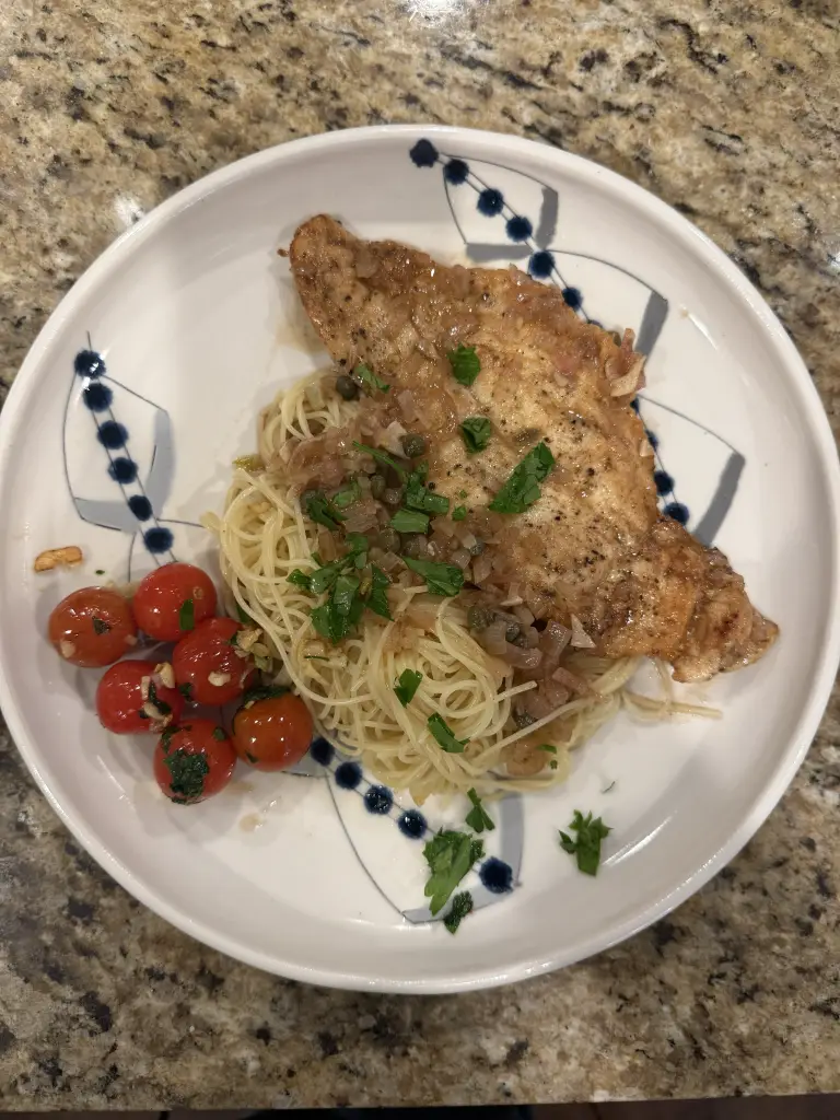 A plate of chicken piccata served over angel hair pasta with a lemon-butter-caper sauce. The chicken is golden and glossy, topped with capers and fresh parsley. On the side are blistered cherry tomatoes, lightly sautéed in butter and garlic. The dish looks bright, fresh, and restaurant-quality.
