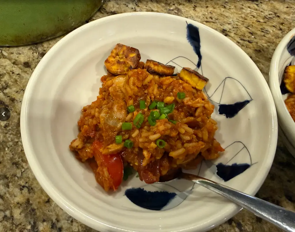 Roasted garlic jollof rice with seasoned chicken thighs and fried plantains served in a bowl with fresh tomatoes and caramelized onions.