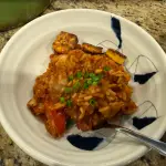 Roasted garlic jollof rice with seasoned chicken thighs and fried plantains served in a bowl with fresh tomatoes and caramelized onions.